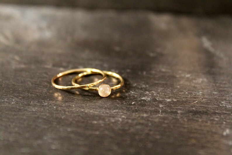 May include: Two gold rings, one with a small, round, light-colored stone, rest on a dark, textured surface. The rings have a simple, elegant design, perfect for everyday wear or special occasions.