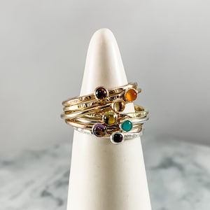 May include: A set of seven gold and silver stacking rings with colorful gemstones. The rings are stacked on a white cone-shaped display stand.