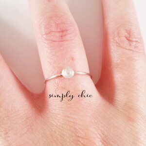 May include: A delicate silver ring with a small, round, white gemstone. The ring is on a finger, with the text "simply chic" in cursive script below the ring.