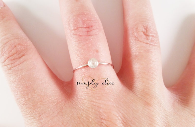 May include: A delicate silver ring with a small, round, white gemstone. The ring is on a finger, with the text "simply chic" in cursive script below the ring.