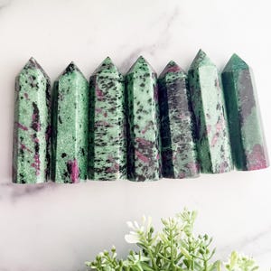 May include: Seven green and pink ruby zoisite crystal towers are arranged in a row. The crystals have a polished, faceted shape with a mix of green and pink hues. The crystals are set against a white marble background with greenery.