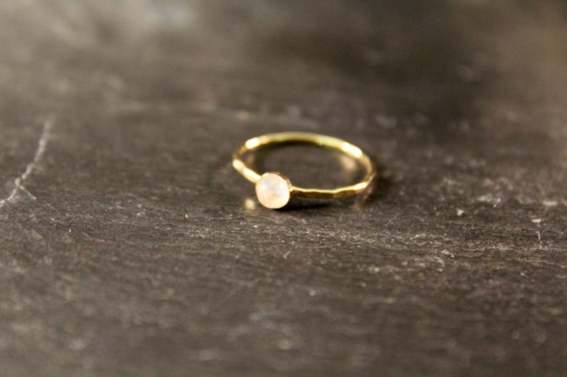 May include: A delicate gold ring with a small, round, pale gemstone. The ring has a hammered texture on the band and sits on a dark, textured surface. The gemstone appears to be a light, neutral color.