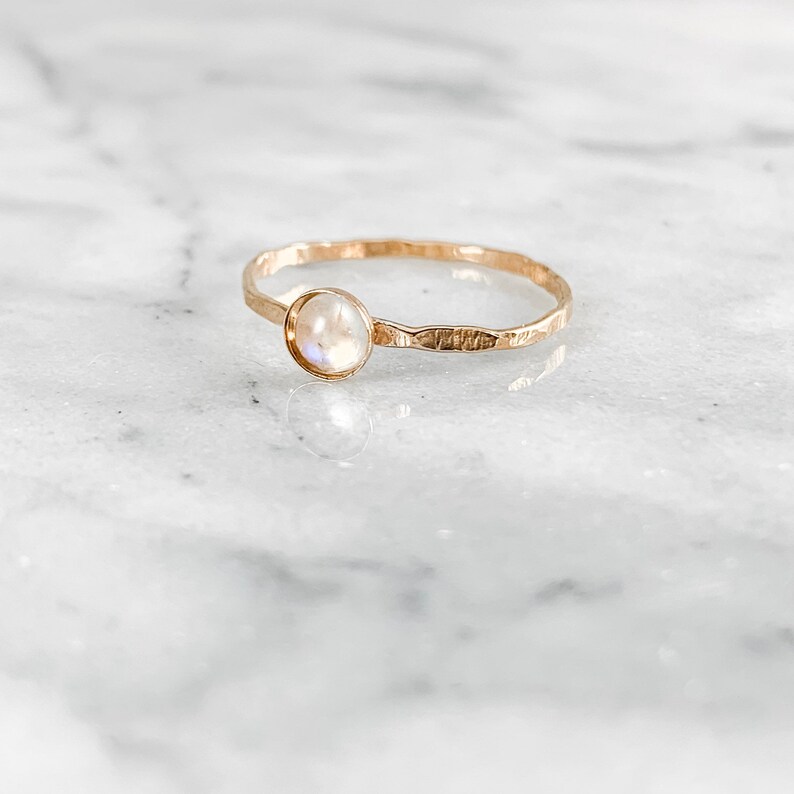 May include: A gold ring with a round, iridescent moonstone. The band has a hammered texture. The ring is set against a white marble surface.