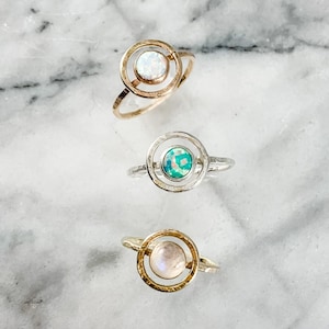 May include: Three rings with circular designs. The top ring is gold-toned with a white opal center. The middle ring is silver-toned with a green opal center. The bottom ring is gold-toned with a light pink opal center. The rings are displayed on a marble surface.