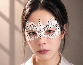 Puede incluir: Una máscara de encaje blanco para baile de máscaras con intrincados patrones florales y pequeños diamantes de imitación negros incrustados en el diseño. La máscara cubre el área de los ojos y presenta un detalle central puntiagudo.