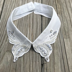 May include: A white, butterfly-shaped collar with intricate beadwork and sparkling rhinestones. The collar is made of a sheer, delicate fabric and features a classic button closure. The design is elegant and suitable for formal wear.