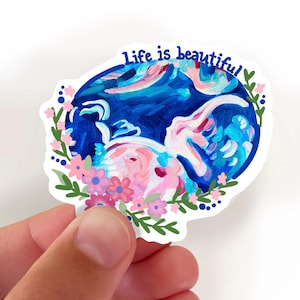 Puede incluir: Una pegatina con una pintura abstracta en tonos azules, rosas y blancos, rodeada de flores rosas y hojas verdes. El texto "Life is beautiful" está escrito en azul en la parte superior.