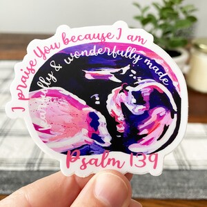 Peut inclure: Un autocollant blanc avec un motif abstrait rose et violet d'un fœtus dans l'utérus. L'autocollant porte le texte "Je te loue parce que je suis merveilleusement et admirablement fait" et "Psaume 139" en rose.
