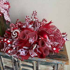 May include: A decorative holiday centerpiece featuring red and white mesh ribbon, candy cane ornaments, and red baubles. The arrangement includes various patterned ribbons with polka dots and stripes, creating a full, textured display.
