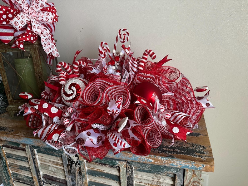 May include: A decorative holiday centerpiece featuring red and white mesh ribbon, candy cane ornaments, and red baubles. The arrangement includes various patterned ribbons with polka dots and stripes, creating a full, textured display.