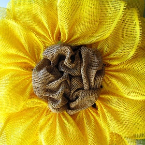 May include: A yellow burlap sunflower wreath with a brown burlap center. The wreath is made of multiple layers of burlap and has a rustic, farmhouse style.