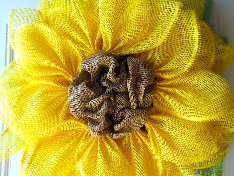 May include: A yellow burlap sunflower wreath with a brown burlap center. The wreath is made of multiple layers of burlap and has a rustic, farmhouse style.