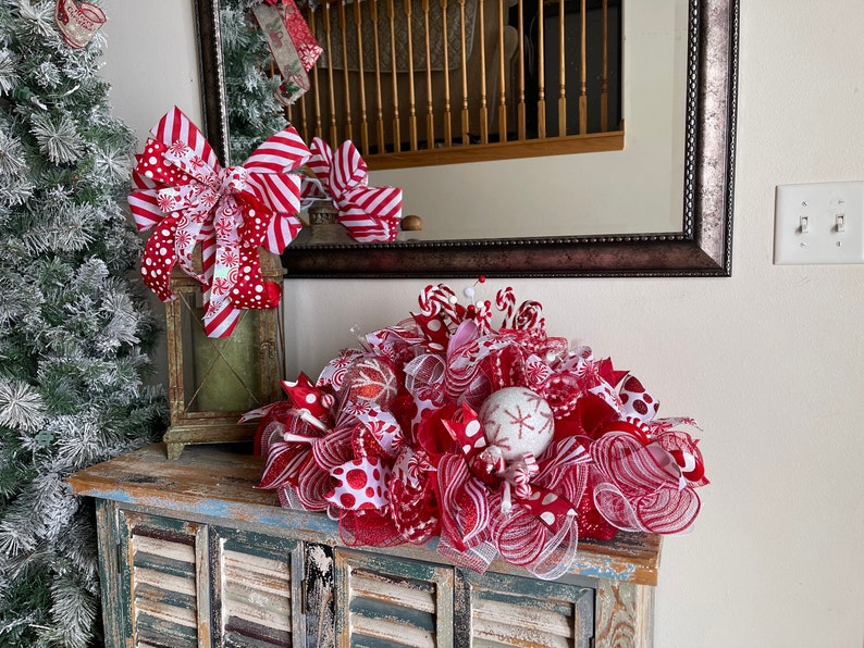 May include: A festive holiday centerpiece featuring red and white mesh ribbon, polka dot patterns, and striped accents. The arrangement includes decorative spheres with snowflake designs and candy-themed elements, creating a layered, textured display.