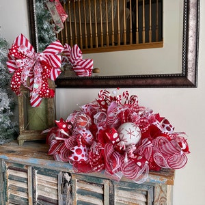 May include: A festive holiday centerpiece featuring red and white mesh ribbon, polka dot patterns, and striped accents. The arrangement includes decorative spheres with snowflake designs and candy-themed elements, creating a layered, textured display.