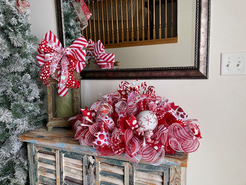 May include: A festive holiday centerpiece featuring red and white mesh ribbon, polka dot patterns, and striped accents. The arrangement includes decorative spheres with snowflake designs and candy-themed elements, creating a layered, textured display.
