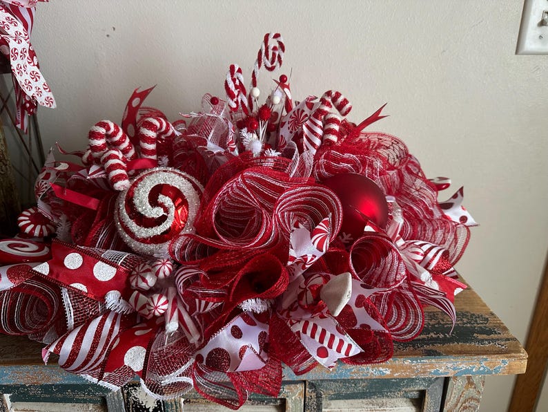 May include: A festive holiday centerpiece featuring red and white mesh ribbon, candy cane ornaments, glittery swirl baubles, and polka dot ribbons. The arrangement is densely packed with layered mesh loops, small peppermint-style accents, and red spheres.
