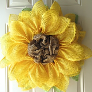 May include: A yellow burlap sunflower wreath with a brown burlap center. The wreath is made of multiple layers of burlap and has a rustic, farmhouse style.