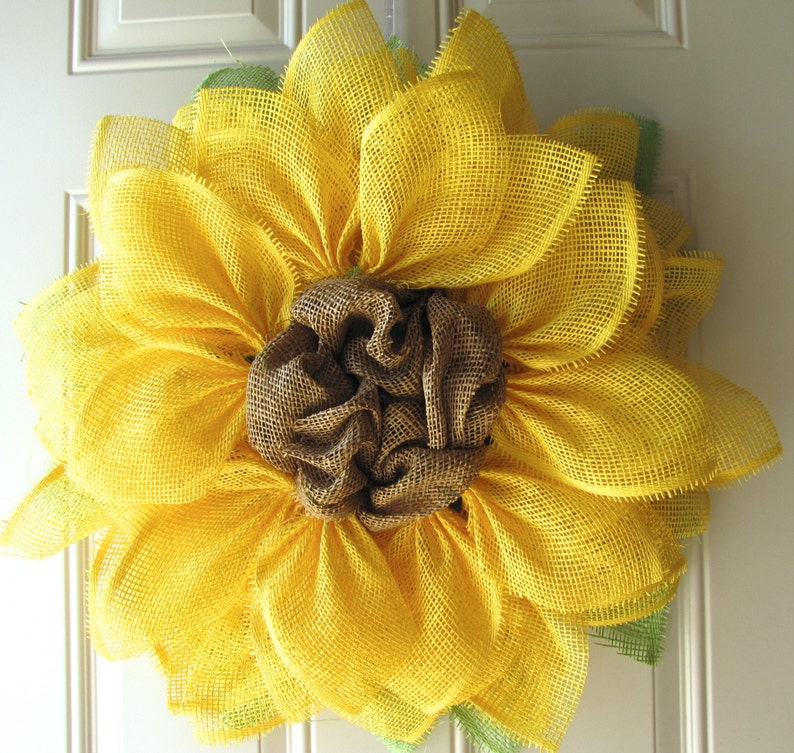 May include: A yellow burlap sunflower wreath with a brown burlap center. The wreath is made of multiple layers of burlap and has a rustic, farmhouse style.