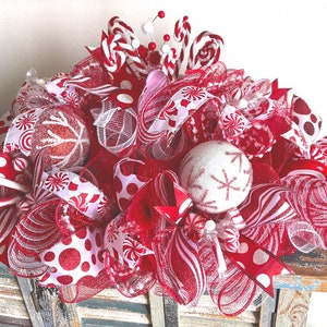 May include: A decorative holiday centerpiece featuring red and white mesh ribbons, glittery polka dot fabric, and round ornaments with snowflake patterns. The arrangement includes candy cane accents and small red and white spheres throughout.