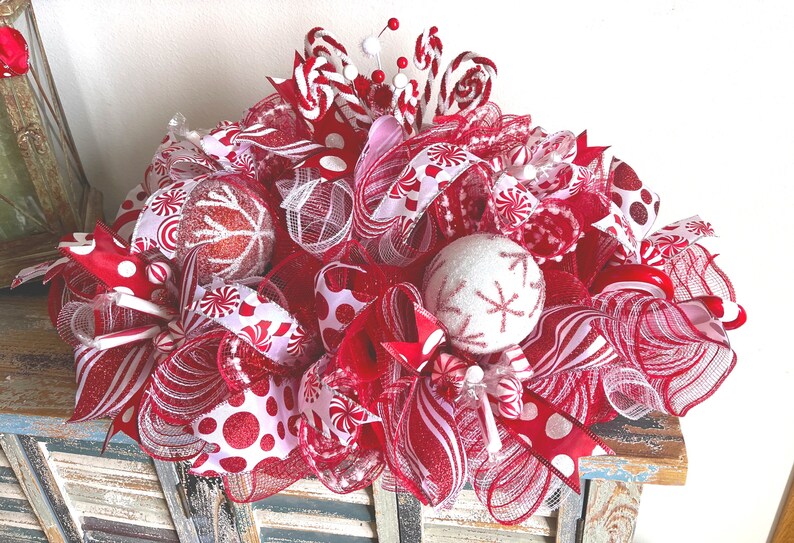 May include: A decorative holiday centerpiece featuring red and white mesh ribbons, glittery polka dot fabric, and round ornaments with snowflake patterns. The arrangement includes candy cane accents and small red and white spheres throughout.