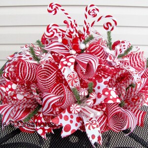 May include: A decorative holiday centerpiece featuring red and white mesh ribbons, polka dot and striped patterns, and peppermint candy accents. The arrangement includes curled wire stems topped with small candy shapes and green pine sprigs.