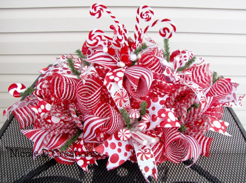 May include: A decorative holiday centerpiece featuring red and white mesh ribbons, polka dot and striped patterns, and peppermint candy accents. The arrangement includes curled wire stems topped with small candy shapes and green pine sprigs.