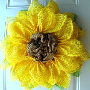 May include: A yellow burlap sunflower wreath with a brown burlap center. The wreath is made of multiple layers of burlap and has a rustic, farmhouse style.