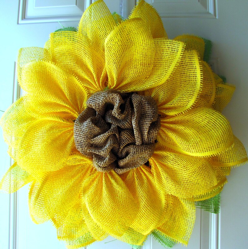 May include: A yellow burlap sunflower wreath with a brown burlap center. The wreath is made of multiple layers of burlap and has a rustic, farmhouse style.