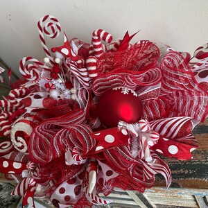 May include: A decorative wreath featuring red and white mesh ribbons, polka dot fabric, and candy-themed accents. The centerpiece is a large matte red ornament with a white snowflake design, surrounded by glittery red and white striped candy canes.
