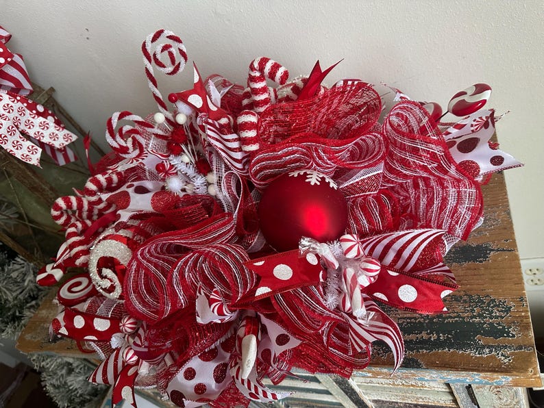 May include: A decorative wreath featuring red and white mesh ribbons, polka dot fabric, and candy-themed accents. The centerpiece is a large matte red ornament with a white snowflake design, surrounded by glittery red and white striped candy canes.