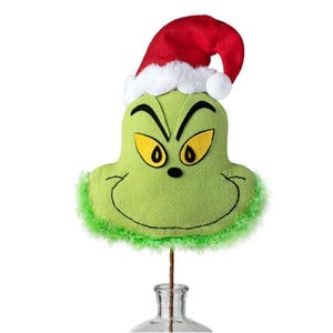 May include: A green plush character head decoration featuring yellow eyes, black eyebrows, and a wide stitched smile. The character wears a red and white felt Santa hat and has a green feathered trim at the base, mounted on a thin brown stick.