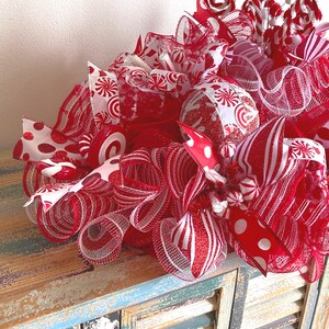 May include: A decorative holiday wreath featuring red and white mesh ribbons, glittery accents, and candy-themed ornaments. The arrangement includes striped patterns, polka dots, and swirling peppermint-style decorations in a festive color palette.