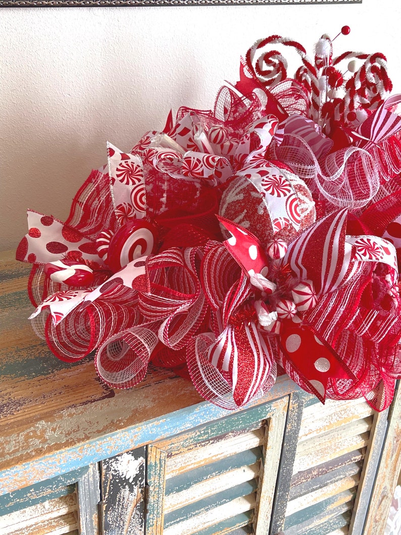 May include: A decorative holiday wreath featuring red and white mesh ribbons, glittery accents, and candy-themed ornaments. The arrangement includes striped patterns, polka dots, and swirling peppermint-style decorations in a festive color palette.
