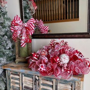 May include: A decorative Christmas centerpiece featuring red and white mesh ribbons, polka dot patterns, striped bows, and glittery ornaments. The arrangement includes small candy cane accents and a white sphere with a red snowflake design.
