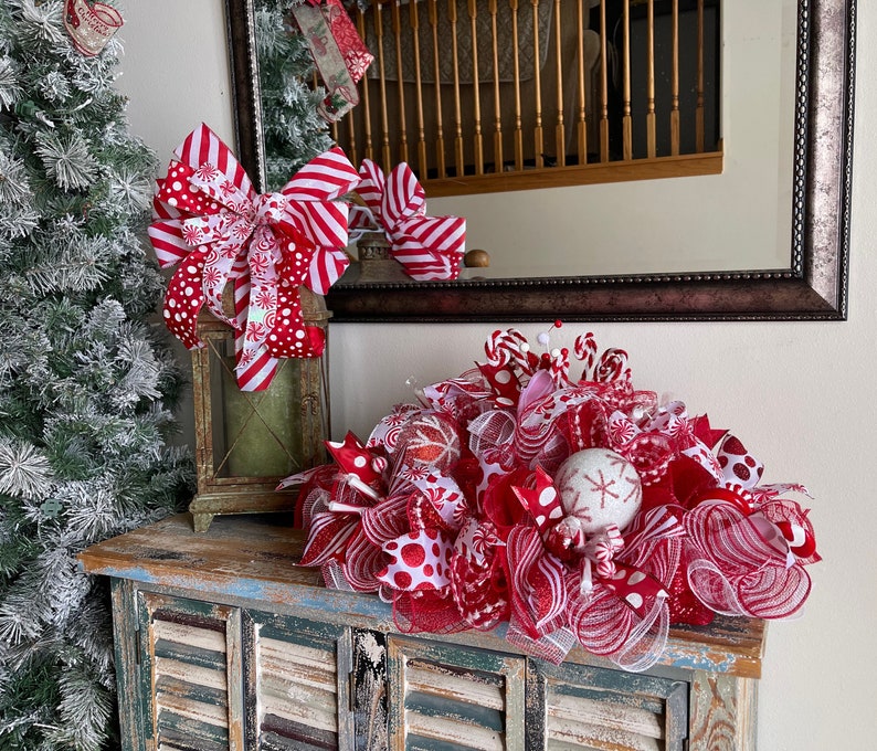 May include: A decorative Christmas centerpiece featuring red and white mesh ribbons, polka dot patterns, striped bows, and glittery ornaments. The arrangement includes small candy cane accents and a white sphere with a red snowflake design.
