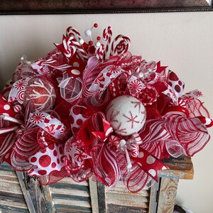May include: A festive holiday centerpiece featuring red and white mesh ribbons, glittery red and white spherical ornaments with snowflake patterns, peppermint candy accents, and small white pom-poms on wire stems arranged in a dense, layered display.