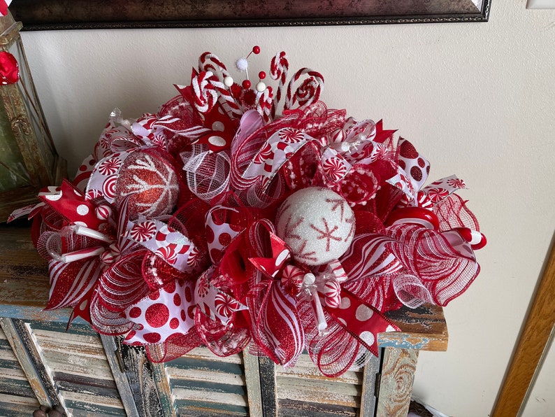 May include: A festive holiday centerpiece featuring red and white mesh ribbons, glittery red and white spherical ornaments with snowflake patterns, peppermint candy accents, and small white pom-poms on wire stems arranged in a dense, layered display.