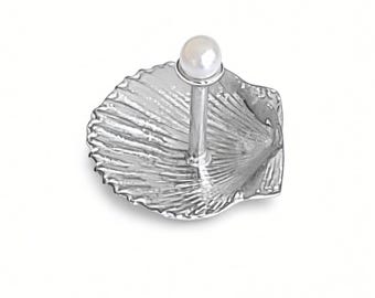 May include: A silver-toned metal ring holder featuring a textured scallop shell base with a vertical post topped by a single, smooth, round white pearl. The object is designed to hold rings on the central post above the shell dish.