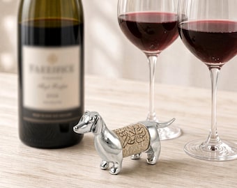 May include: A silver-colored metal dachshund figurine bottle stopper featuring a central cork body section with an engraved grape vine and leaf design. The dog-shaped stopper has a smooth, polished finish on the head, legs, and tail.