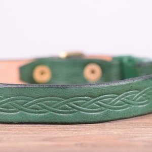 May include: A green leather bracelet featuring a Celtic knot pattern. The bracelet has a brass buckle and is approximately 2 cm wide. The inside of the bracelet is a natural tan colour.