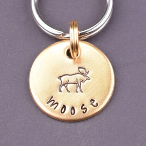 Peut inclure: Porte-clés en métal doré avec une étiquette ronde représentant une silhouette d'élan et le mot "moose" gravé dessus. Le porte-clés est attaché à un anneau en métal argenté.