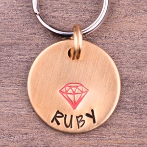 May include: A round gold-coloured dog tag with a red diamond and the name "RUBY" engraved on it. The tag is attached to a silver key ring.