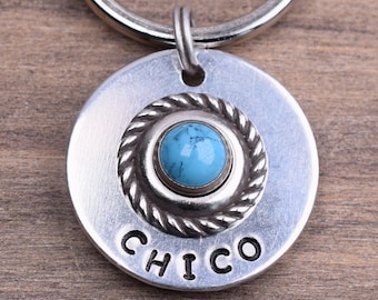 Op de afbeelding: Zilveren sleutelhanger met een rond label met de tekst "CHICO" en een turquoise steen in een zilveren zetting.