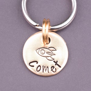 以下が含まれることがあります： 銀色のキーリングが付いた金色のキーホルダー。キーホルダーには、ロケットと「コメット」という文字が刻まれた丸いタグが付いています。