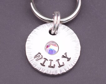 May include: Silver dog tag engraved with the name "WILLY". The tag features a small iridescent crystal in the centre and is attached to a silver key ring.