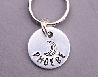 May include: A circular silver-toned metal keyring tag featuring a stamped crescent moon symbol above the name PHOEBE in capital letters. The tag is attached to a standard silver-toned metal split ring, providing a functional and simple design.