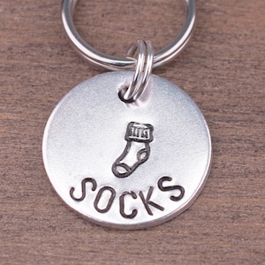 May include: A circular, silver-coloured keyring tag featuring a stamped sock graphic and the word "SOCKS" in capital letters. The tag is attached to a silver split ring. The background is a brown wooden surface.