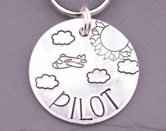 May include: Silver keychain with a round tag featuring a plane, clouds, and a sun. The tag also says "PILOT" in a stylized font.