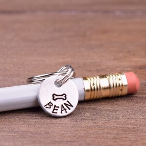 Könnte beinhalten: Silbernes Hundeanhänger mit einem Knochen und dem Namen "BEAN" eingraviert. Der Anhänger ist an einem silbernen Schlüsselring befestigt und liegt auf einem weißen Bleistift.