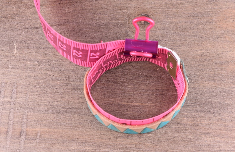 May include: Pink measuring tape wrapped around a brown leather bracelet with a turquoise and brown pattern. The tape is held together with a purple binder clip.